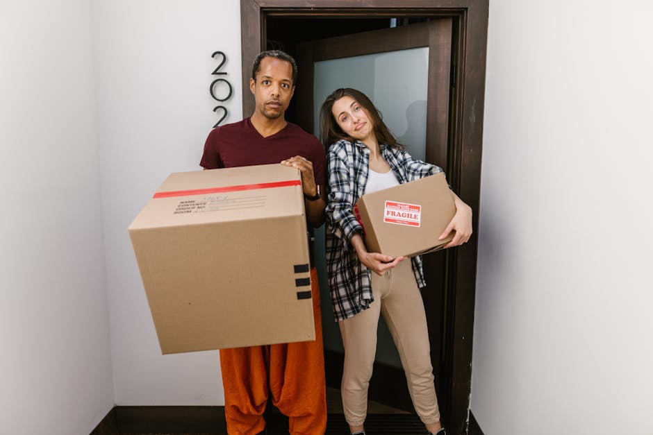 A man and woman engaged in a home relocation process outside a residential property, facing each other with the woman presenting a cardboard box marked 'Fragile' with a red and white warning label, wh